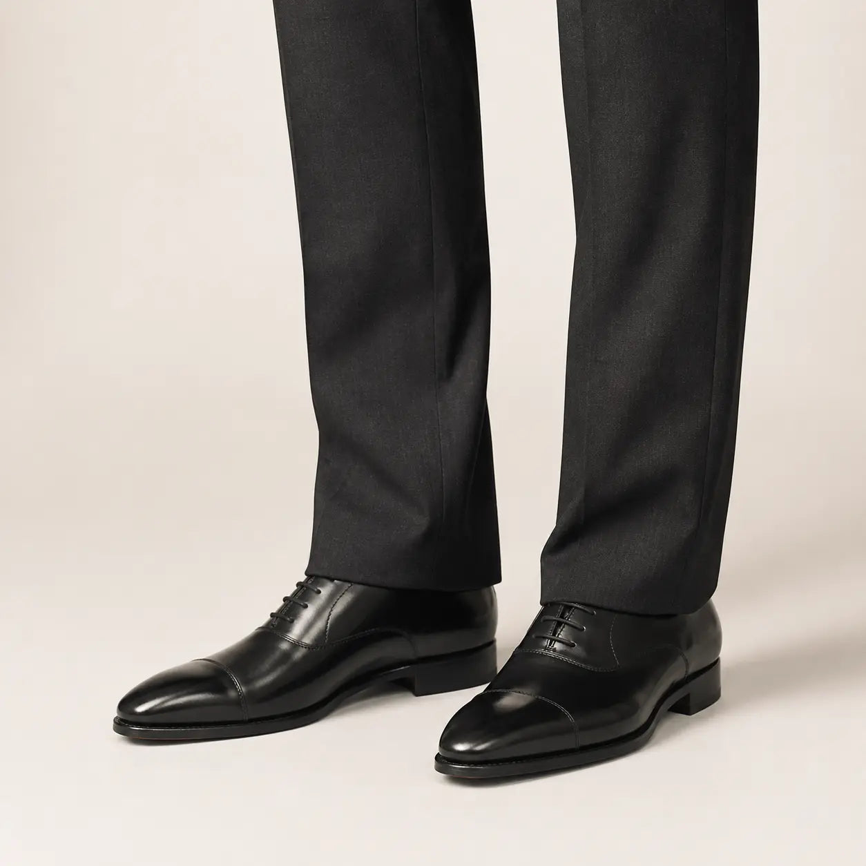 Black cap-toe Oxford shoes worn with charcoal tailored trousers in a warm studio.