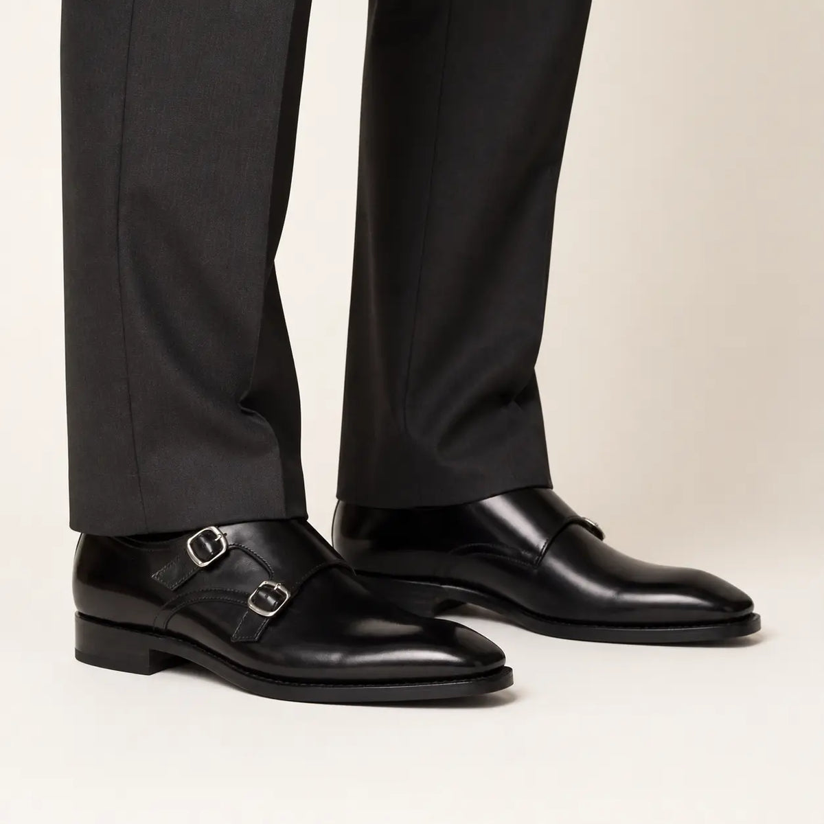 Black double monk strap shoes worn with charcoal tailored trousers in a warm studio.