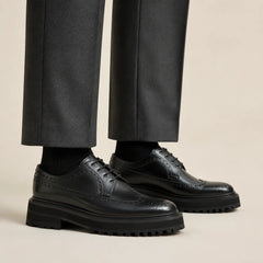 Elongated black wingtip brogue Derby shoes with black lug soles worn with separate charcoal trousers and black socks