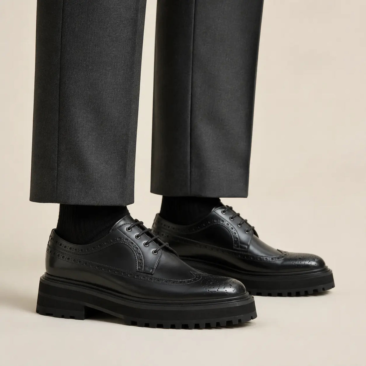 Elongated black wingtip brogue Derby shoes with black lug soles worn with separate charcoal trousers and black socks