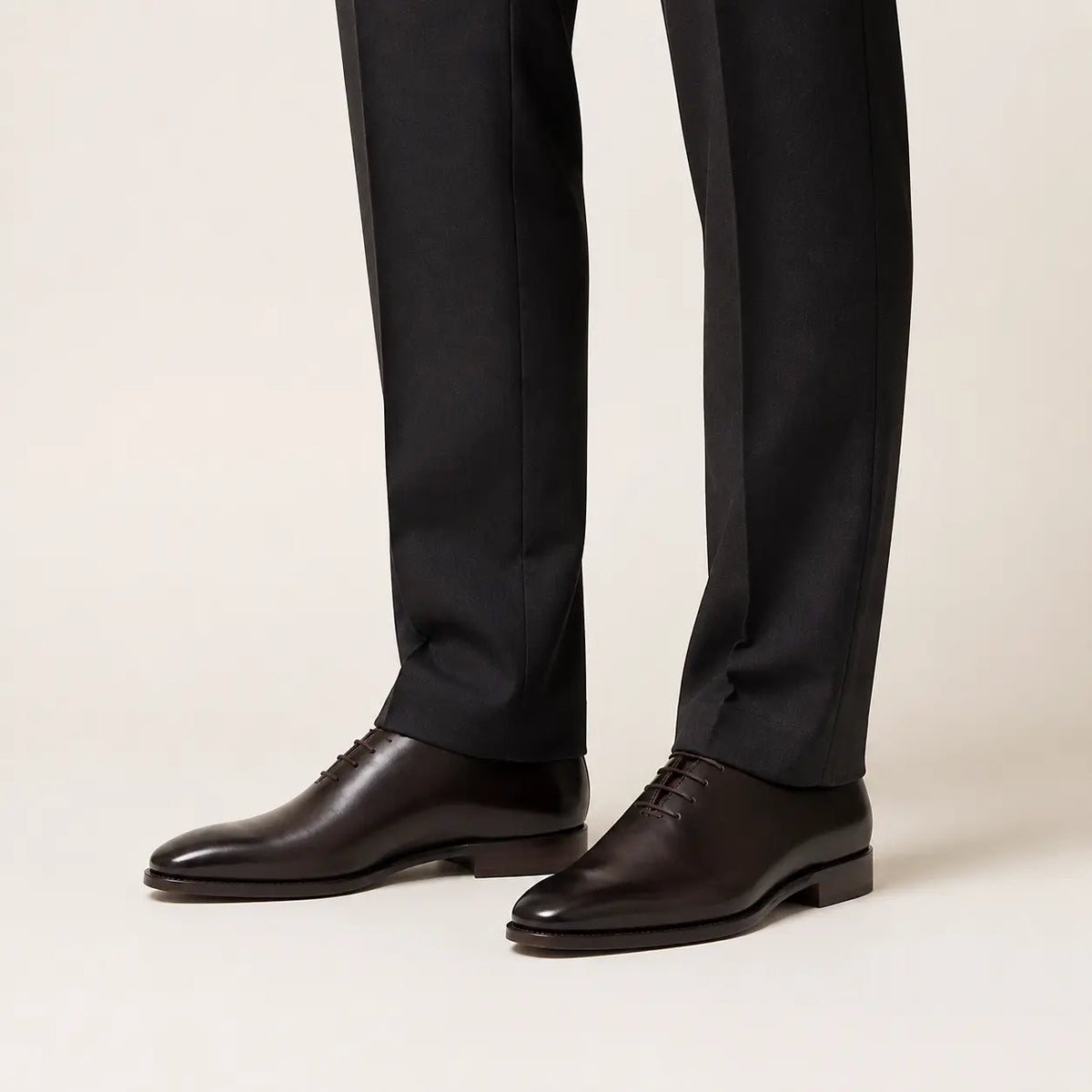 Dark brown wholecut Oxford shoes worn with charcoal tailored trousers in a warm studio.
