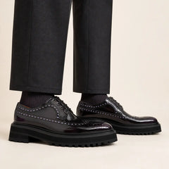 Elongated polished dark plum brogue Derby shoes with black lug soles worn with relaxed straight charcoal trousers and dark socks